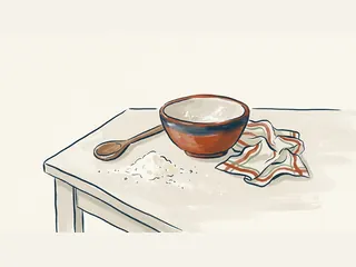 Cuenco de cerámica, cuchara de madera, un poco de harina derramada y un paño sobre una mesa.