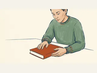 Una persona sostiene un libro cerrado de cubierta lisa sobre una mesa.