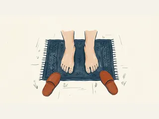 Dos pies descalzos sobre una alfombra azul, con unas zapatillas al lado.